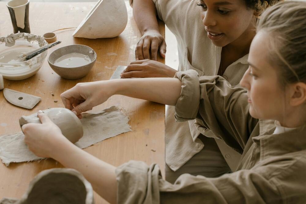 Två personer arbetar med keramik vid ett träbord, formar lera med händerna.