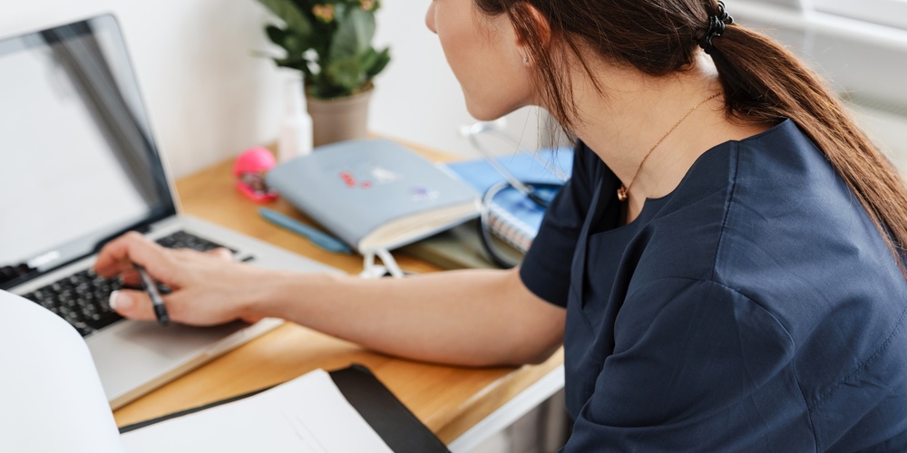 Kvinna i blå arbetskläder arbetar vid skrivbord med laptop och papper, sedd bakifrån.