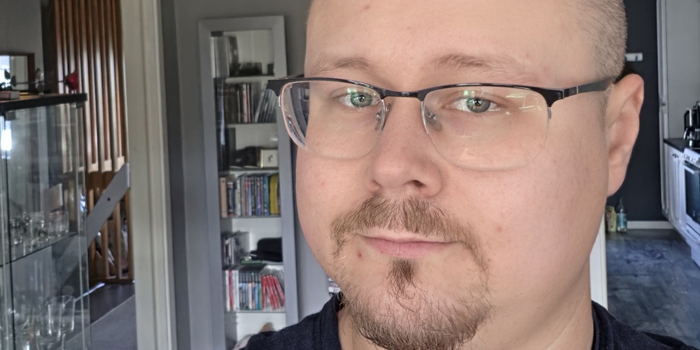 Man med glasögon och kort skägg tar en selfie i ett modernt vardagsrum med grå väggar.