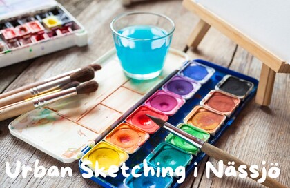 Akvarellfärger, penslar och ett glas vatten på ett träbord med texten Urban Sketching i Nässjö.