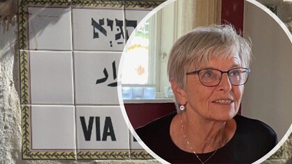 Äldre kvinna med glasögon och kort grått hår sitter inomhus. Gatuskylt med texten 'VIA' syns till vänster.