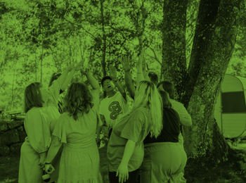 En grupp människor står i en ring utomhus och sträcker upp händerna mot mitten under träd.