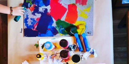 Ett bord med färgglada konstmaterial, papper, färg, penslar och två barns händer som skapar konstverk.