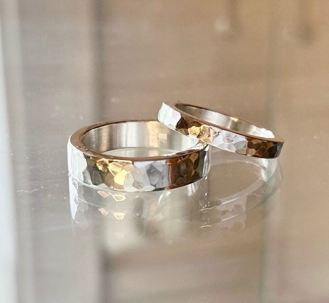 Två ringar i silver och guld med hamrad yta ligger på en reflekterande yta.