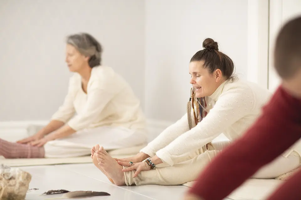 Två kvinnor sitter på golvet i ett ljust rum och gör sittande yogaövningar.