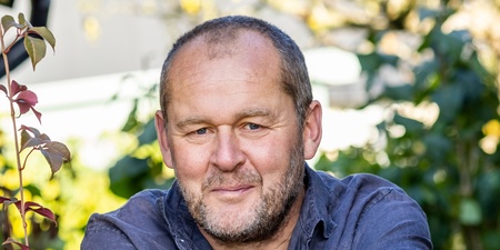 En person står i en trädgård med armarna vilande på ett trädgårdsredskap, omgiven av gröna växter.