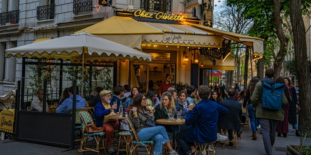 Uteservering på ett franskt café med människor som samtalar och äter under en gul markis.