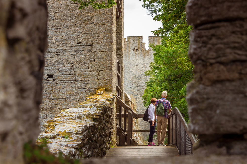 Två personer med ryggsäckar promenerar längs en gammal stenmur, omgivna av grönska.