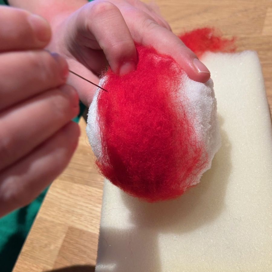 En person nålfiltar röd ull på en vit boll ovanpå en skumgummibit på ett träbord.