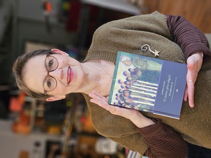 Kvinna med glasögon håller upp en bok och ler mot kameran i en inomhusmiljö.