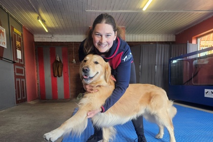En kvinna ler och håller en glad golden retriever på ett blått golv i ett inomhusutrymme.