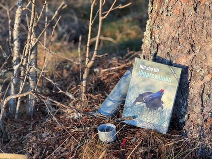 En bok med en fågel på omslaget, en termos och en kopp står lutade mot ett träd i skogen.