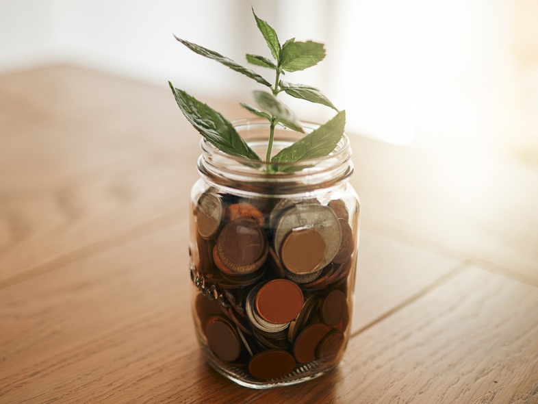 En glasburk fylld med mynt står på ett träbord, med en grön växt som växer ur burken.