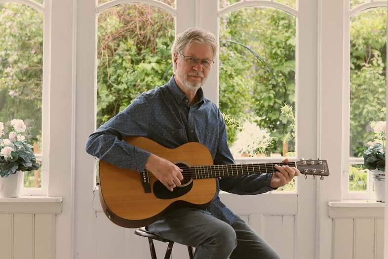 Person sitter på en stol och spelar akustisk gitarr i ett ljust rum med stora fönster och blommor.