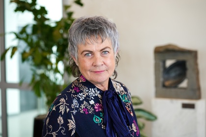 Äldre kvinna med grått hår i blommig kofta står inomhus med växter och konst i bakgrunden.