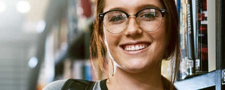Ung kvinna med glasögon och ryggsäck håller böcker och ler i ett bibliotek.