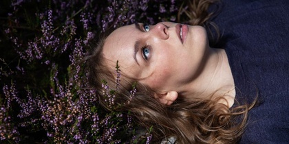 Kvinna med blå ögon ligger på rygg bland lila blommor och tittar uppåt.