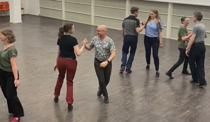 Sex personer dansar pardans i en ljus sal med trägolv.