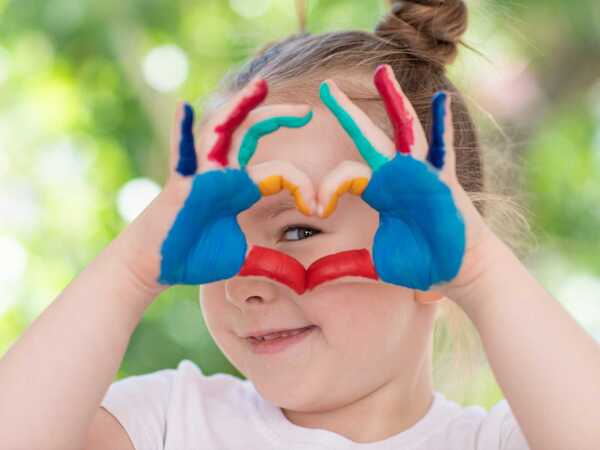 Ett barn håller upp händerna och formar ett hjärta. Barnet har målade fingrar och handflator och kikar med ena ögat genom hjärtat.