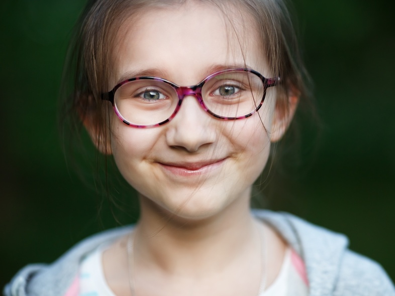 En ung flicka med glasögon ler mot kameran, suddig grön bakgrund.