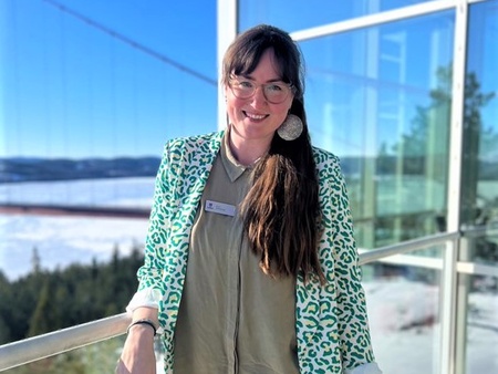 Person med långt hår och glasögon står leende på en solig balkong med vinterlandskap i bakgrunden.