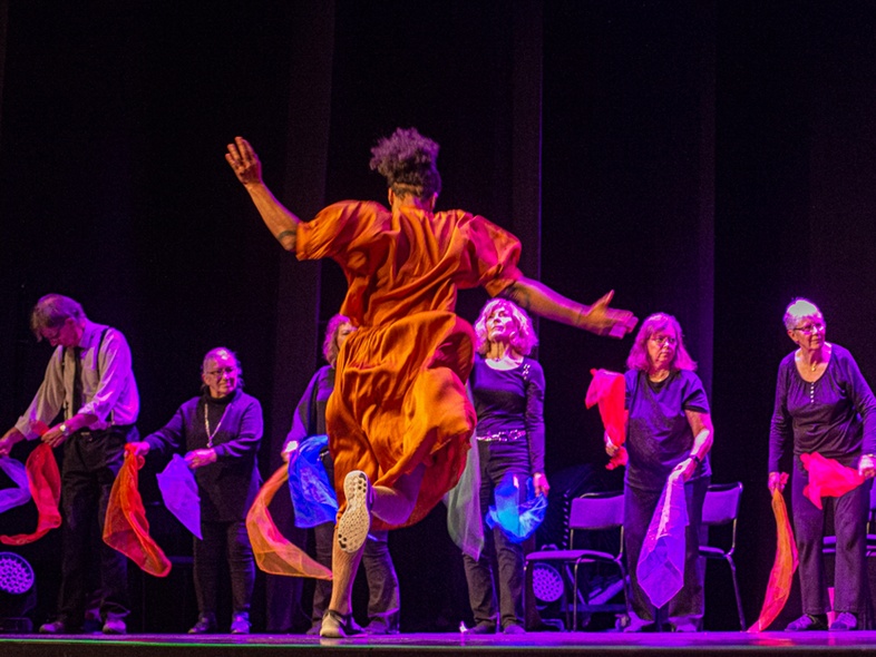 En person i orange klänning dansar framför en grupp äldre personer med färgglada sjalar på en scen.
