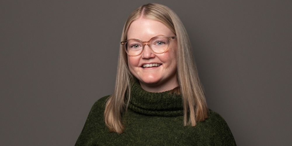 Person med långt blont hår, grön stickad tröja och svarta byxor står mot en grå bakgrund.