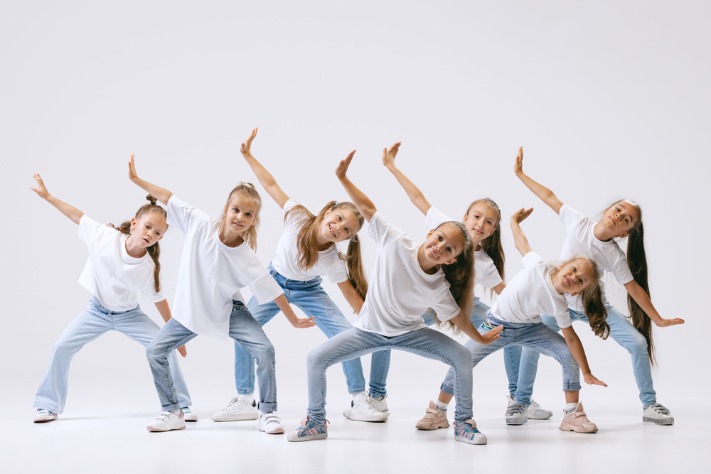 Sju barn i vita t-shirts och jeans dansar tillsammans i en grupp mot vit bakgrund.