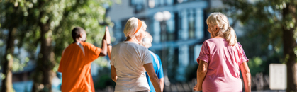 Fyra äldre personer promenerar utomhus i en park med träd och byggnader i bakgrunden.