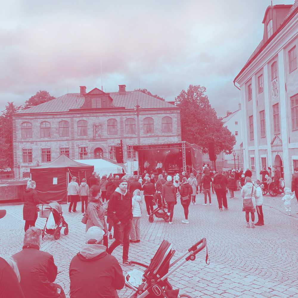 En folksamling på ett torg framför en scen med livemusik, omgiven av gamla stenbyggnader.