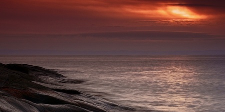 Solnedgång över ett lugnt hav med släta klippor i förgrunden och dramatiska moln på himlen.