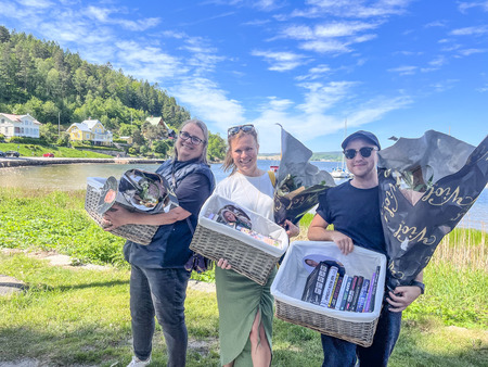 Tre personer står utomhus med presentkorgar och blommor, leende mot kameran vid en sjö och grönska.