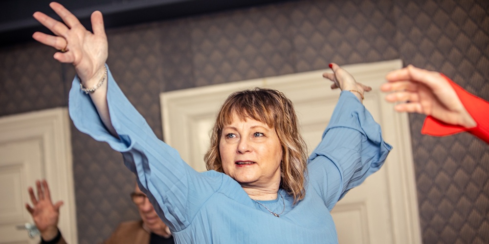 Kvinna i blå blus sträcker ut armarna och deltar i en gruppaktivitet inomhus.