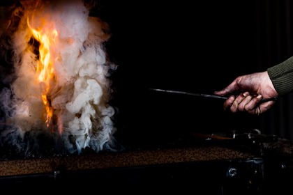 En hand håller ett metallverktyg nära en eld med tjock vit rök mot svart bakgrund.