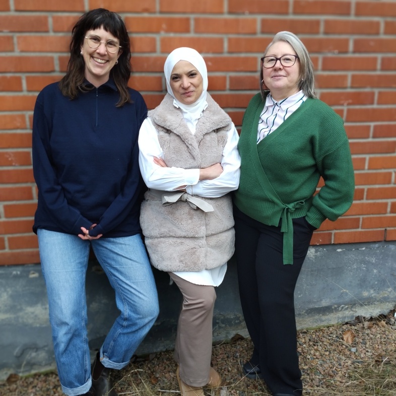 Tre personer står framför en tegelvägg utomhus, klädda i vardagskläder.