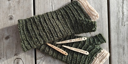 Två gröna, ribbstickade fingerlösa vantar med ljusbeige kontrastkant, fotograferade uppifrån på ett underlag av grå träpanel. Ovanpå vantarna ligger tre smala, handdekorerade träknappar med inbrända dekorativa mönster
