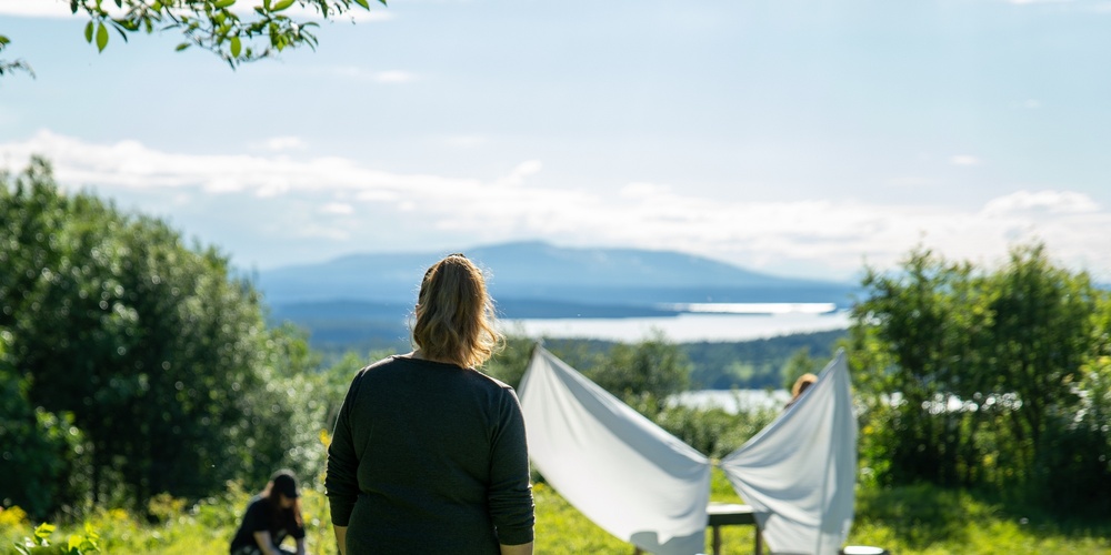 En person står på en gräsmatta och tittar mot ett tält av vita lakan, med fler personer i bakgrunden och sjöutsikt.