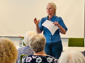 En kvinna håller en presentation framför en grupp äldre personer i ett mötesrum med gröna stolar.