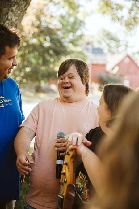 Tre personer står utomhus och skrattar tillsammans, en håller i en mikrofon och en annan i en gitarr.