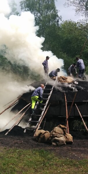 Flera personer arbetar på en rykande kolmila utomhus omgiven av träd och säckar.