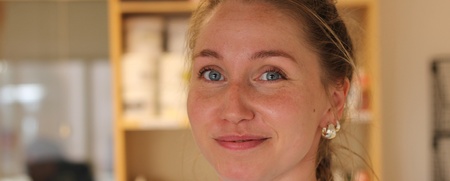 Kvinna med blont hår och blå ögon ler mot kameran i en inomhusmiljö.