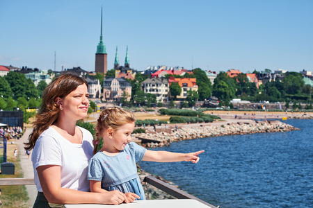 En kvinna och ett barn står vid vattnet och tittar ut över en stad i solsken.