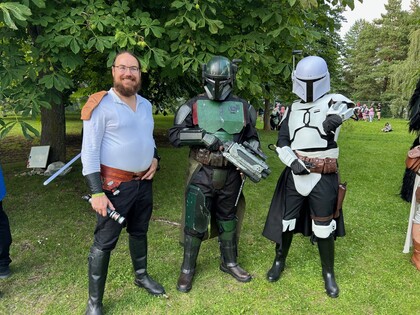 Tre personer i Star Wars-inspirerade dräkter poserar utomhus på gräs, två i Mandalorian-rustningar.