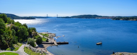 Solig badplats vid havet med gräsytor, klippor och en bro i bakgrunden.