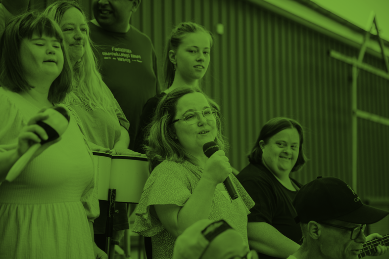 En grupp människor, varav en kvinna håller en mikrofon, sitter tillsammans inomhus. Bilden har grön färgton.