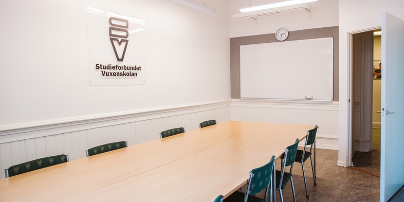 Ett tomt konferensrum med långbord, gröna stolar, whiteboard och klocka på väggen.