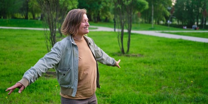 Person står med utsträckta armar i en grön park, ser glad ut och njuter av naturen.