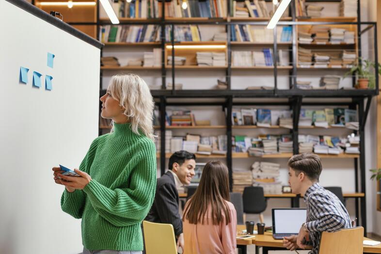 Kvinna i grön tröja sätter post-it-lappar på whiteboard medan tre personer sitter vid ett bord i ett bibliotek.