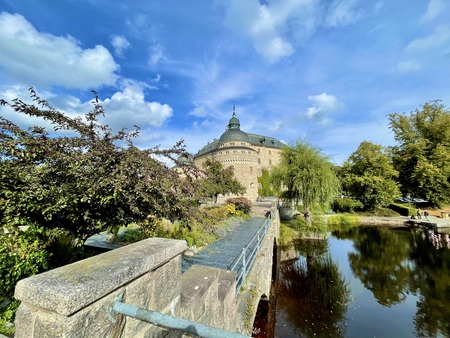 En gammal borg med rund tornstruktur, omgiven av grönska och vatten under en blå himmel med moln.