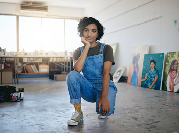 Ung kvinna i blå snickarbyxor sitter på huk i en konststudio med målningar i bakgrunden.